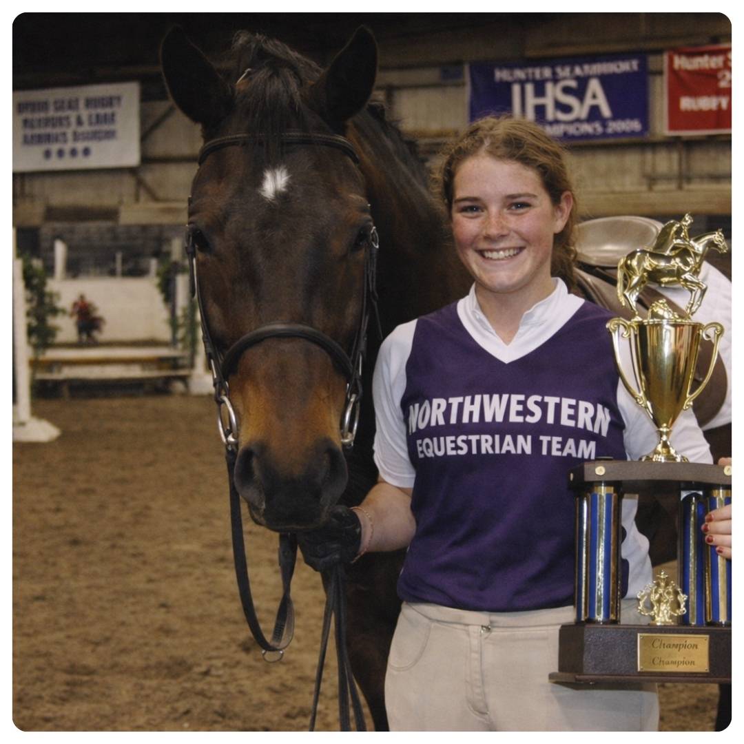 Woman in Northwestern Equestrian Team shirt holding a horse and trophies in an equestrian arena.